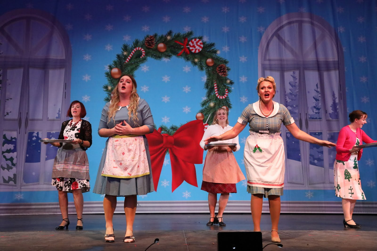 Five women dance in front of a indoor holiday scene.