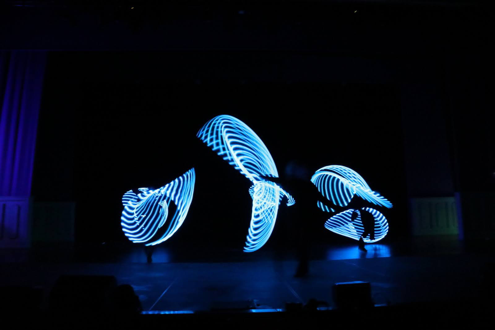 Three dancers with illuminated capes on a darkened stage.